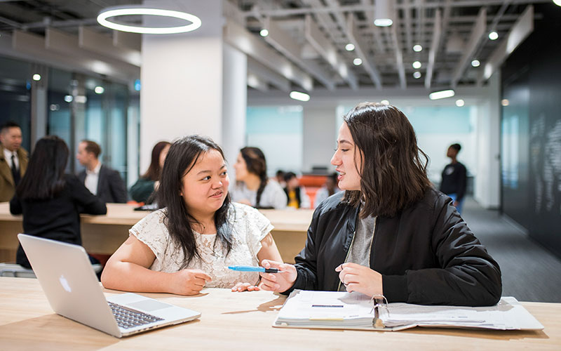 Two international students are studying and talking to each other on campus. Other students can be seen in the background.