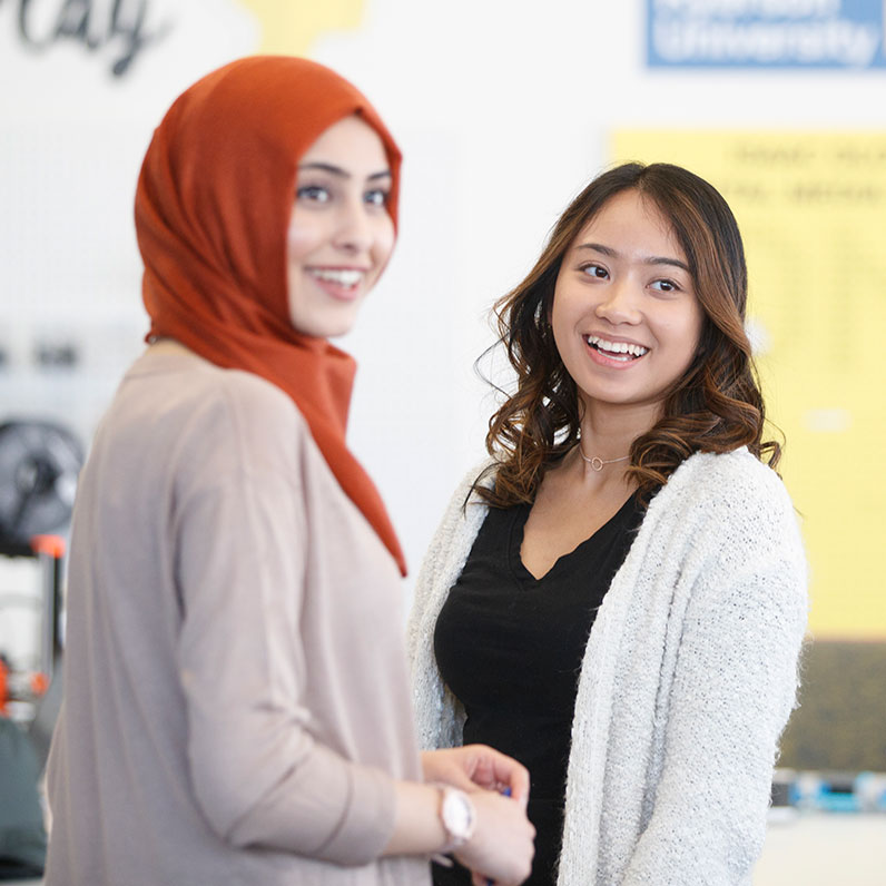 Two female students standing together, looking at someone off camera and smiling.