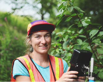 Geography Researcher, Claire Oswald, standing in a forest wearing a safety vest. 