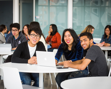 A group of students sits around a Mac laptop at a table. 