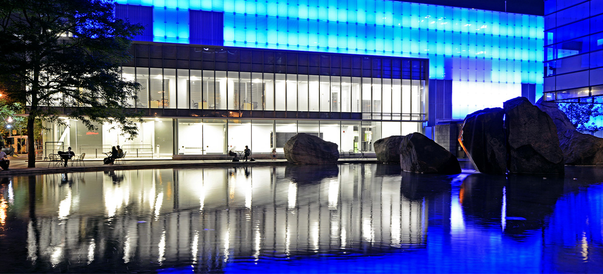 Water feature on campus with large rocks interspersed on one end in front of a brightly lit building with people sitting in front of it.