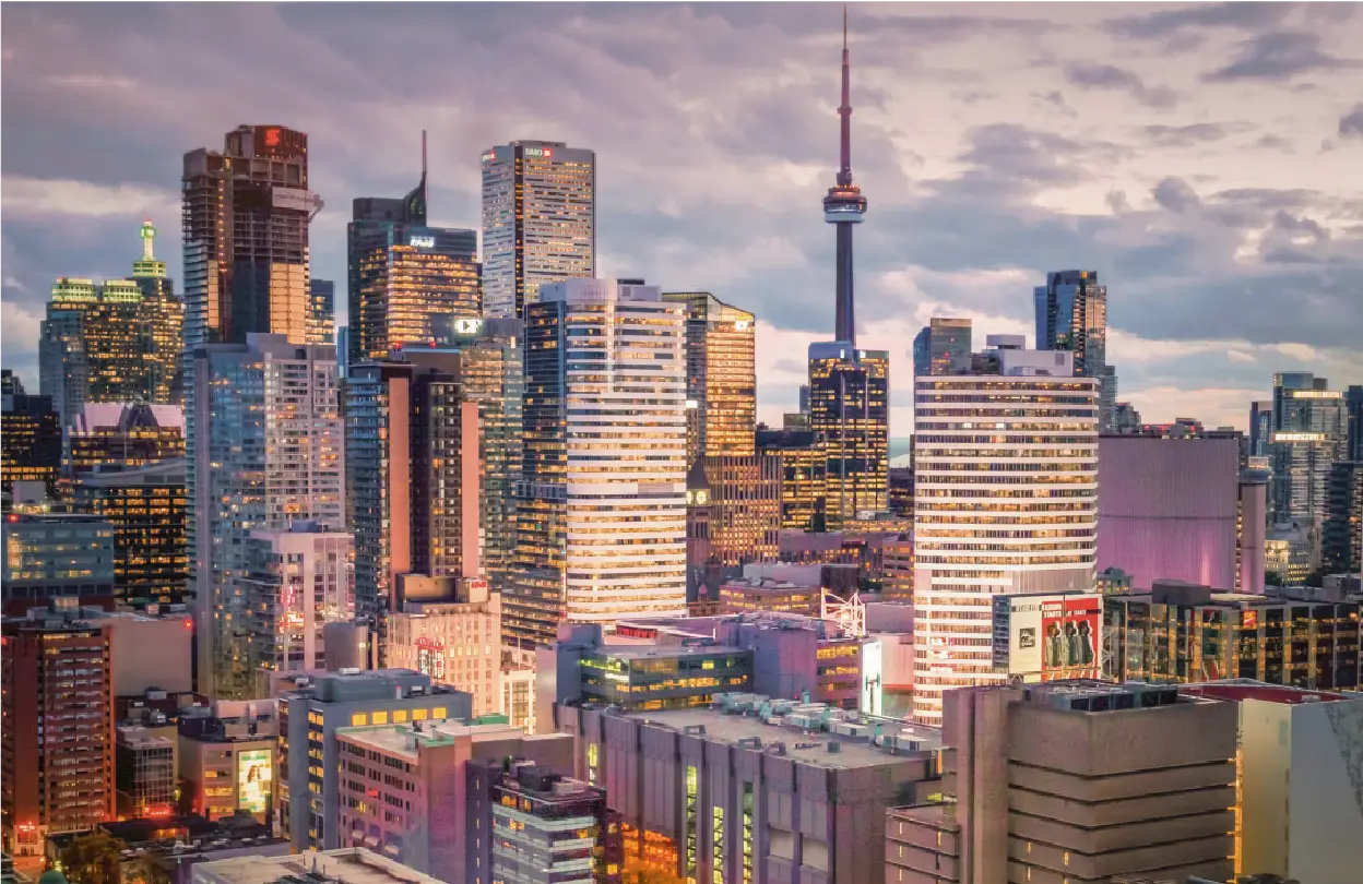 Early evening photo of the Toronto cityscape with the CN Tower in the middle and well-lit buildings surrounding it against a cloudy sky.
