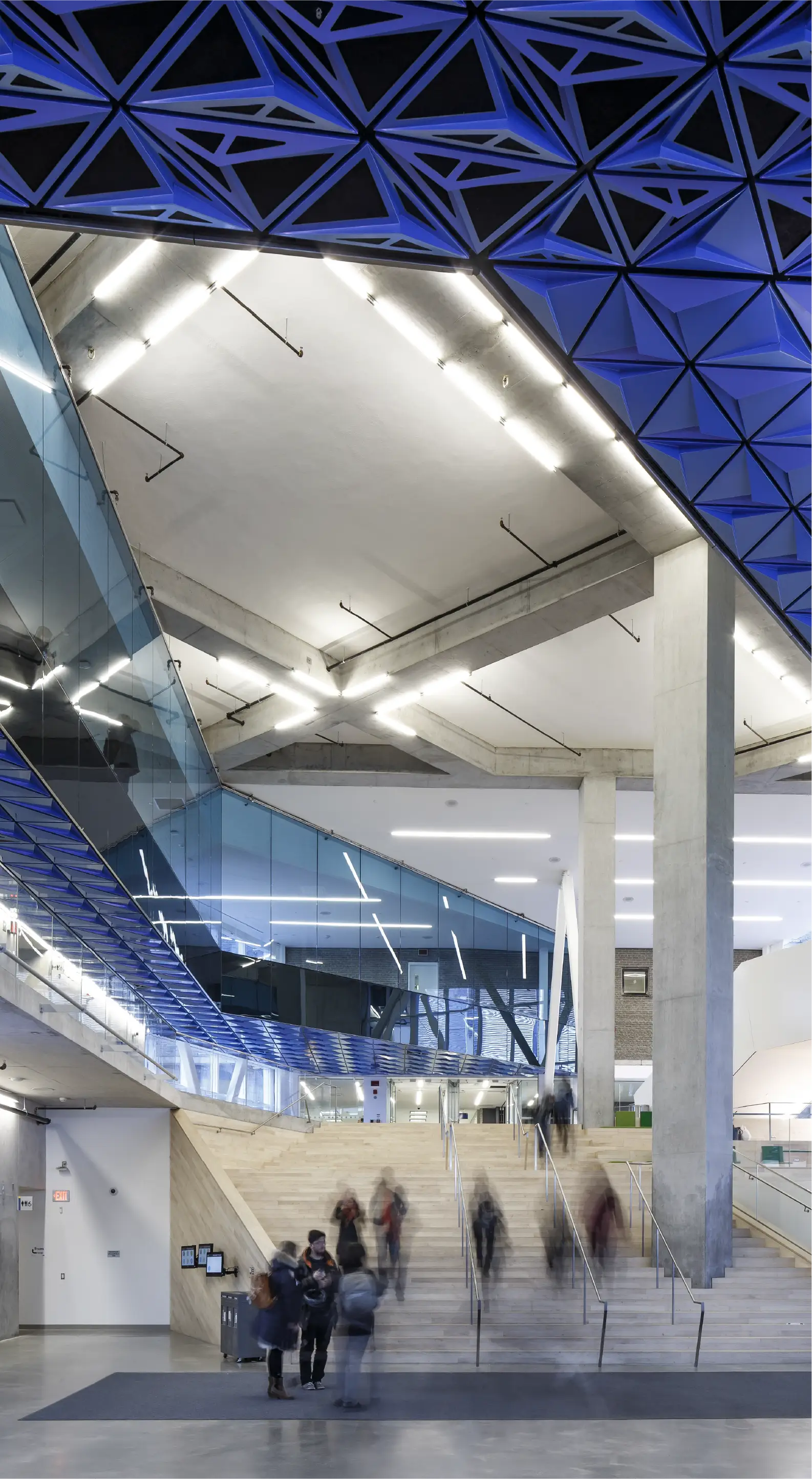 Indoor view from lobby to ceiling of SLC building with blurry motion image of people descending stairs and standing at base of stairs.