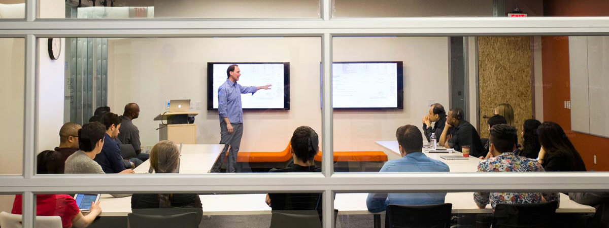 A teacher instructs a small class of students, as viewed through a window to the room