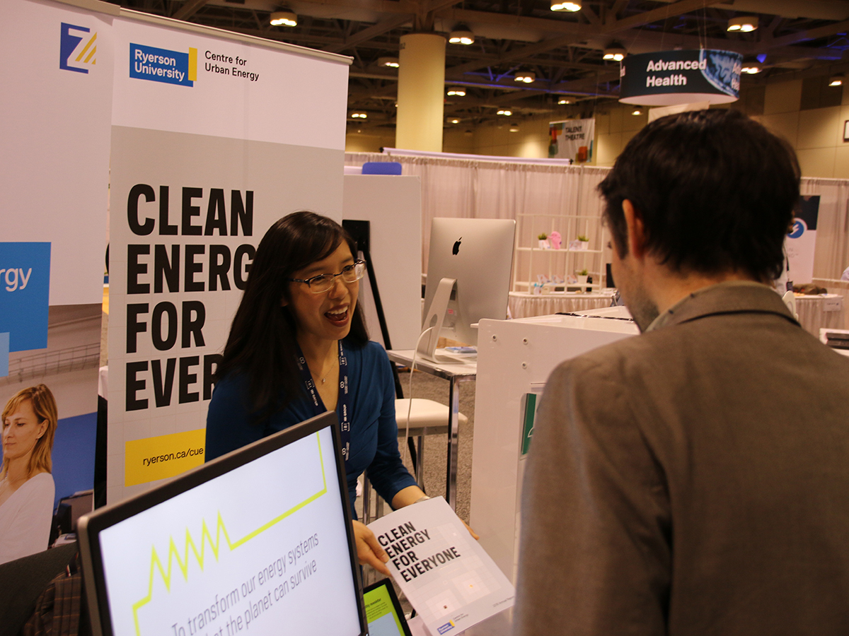 In front of a banner that reads "Clean Energy for Everyone", a woman hands an OCR Discovery attendee a pamphlet