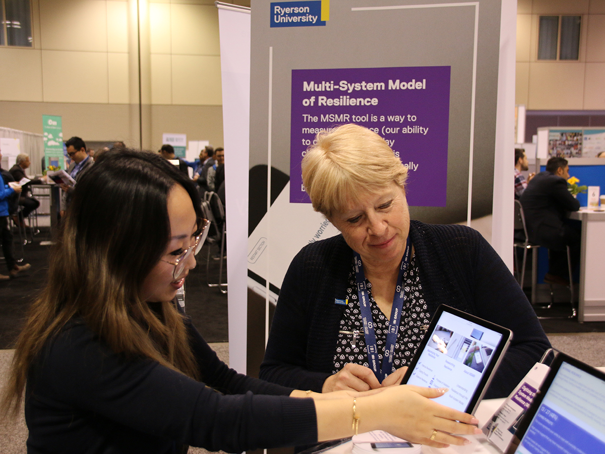 In front of a banner that reads "Multi-System Model of Resilience", two women read information off of tablet computers
