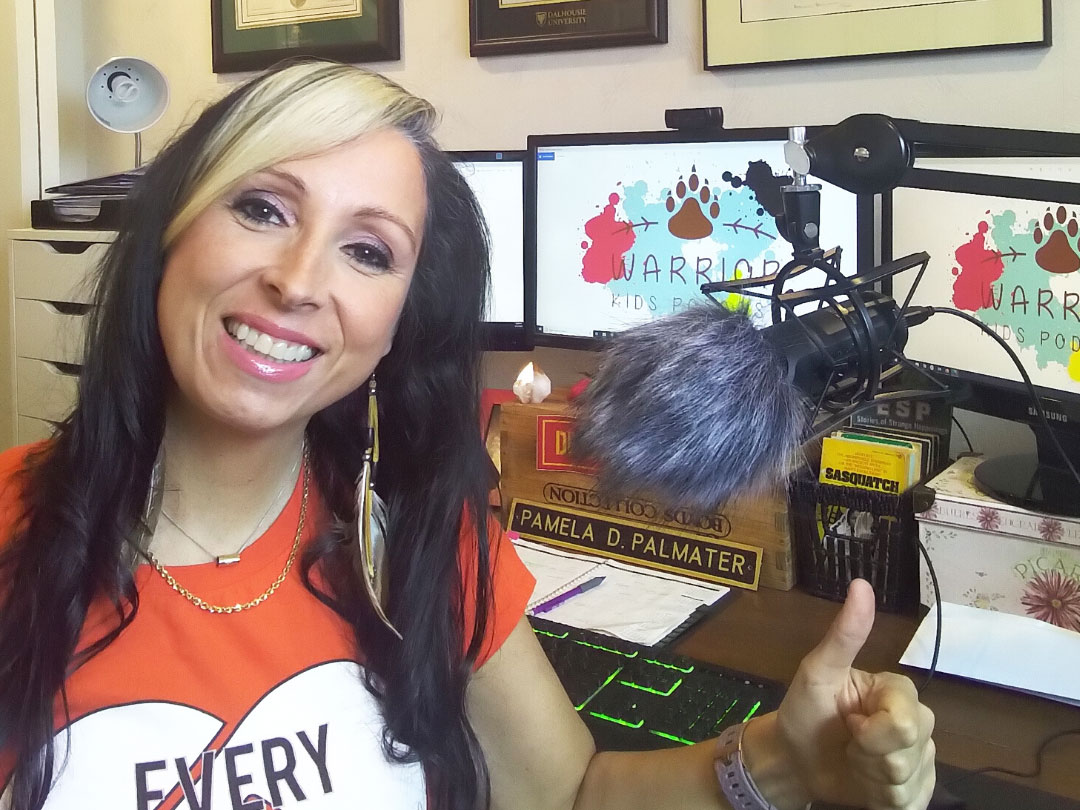 Photo of professor Pamela Palmater, a Mi’kmaw woman with blonde and black hair. She is wearing feather earrings and an orange shirt. Behind her are computer screens and a microphone, which she uses to record podcasts.