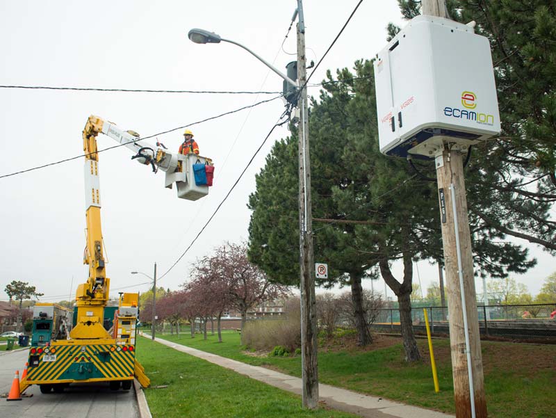 A man in an orange jacket and yellow hardhat is elevated in a box by a machine to work on nearby power lines.