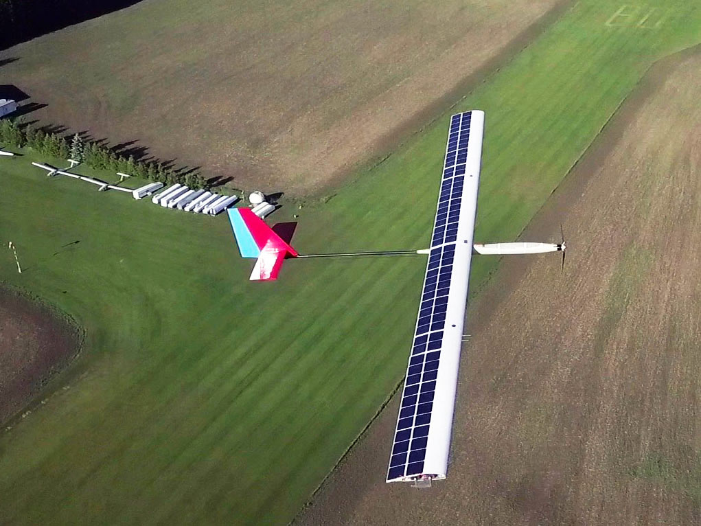 A small, lightweight, unmanned aircraft with solar panels on its wings in mid-flight over farm fields and homes.