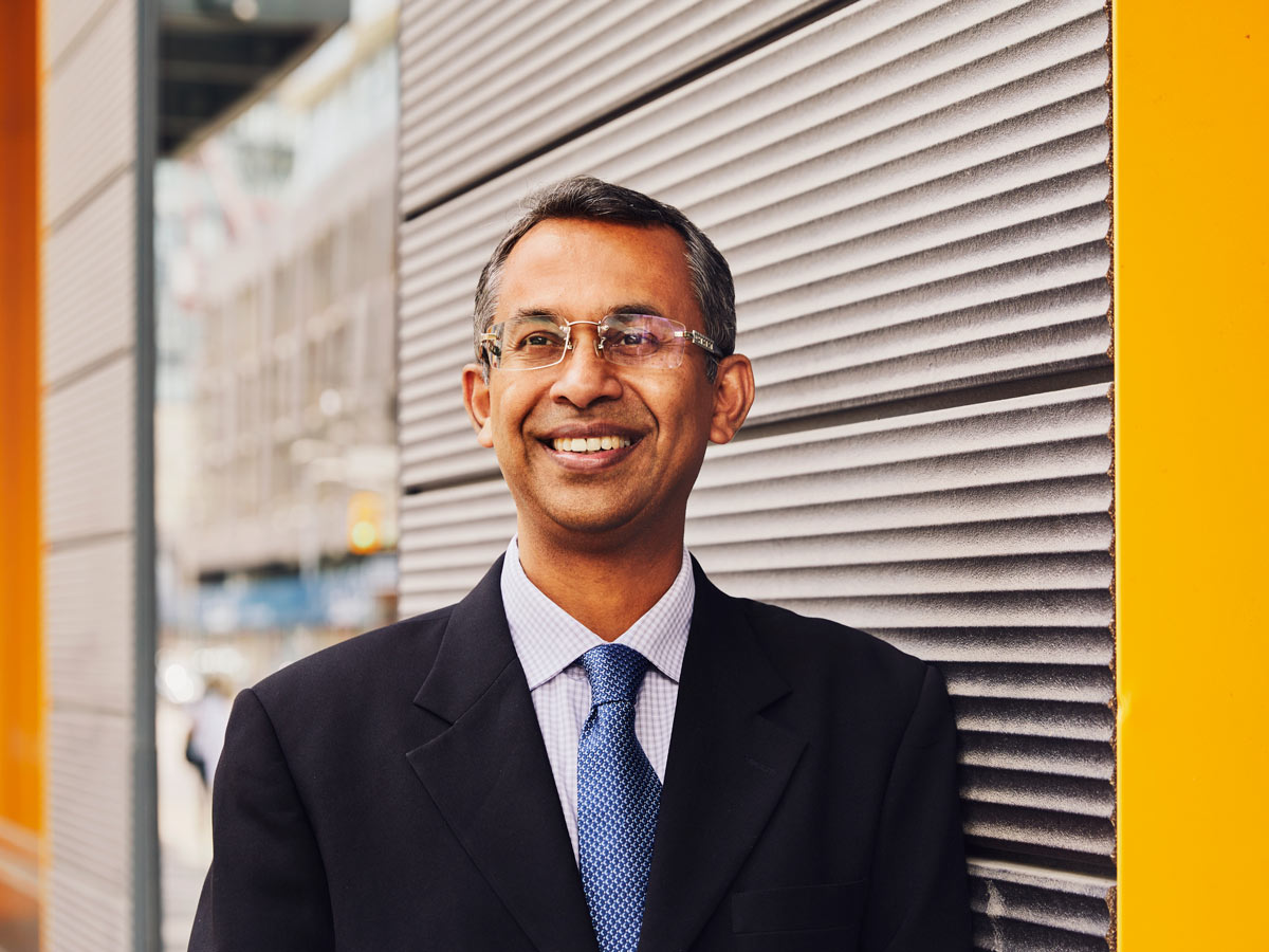 Professor Bala Venkatesh, wearing rimless glasses, a white and blue checked shirt and a blue tie under a black blazer, smiles as he stands against a corrugated steel wall. 