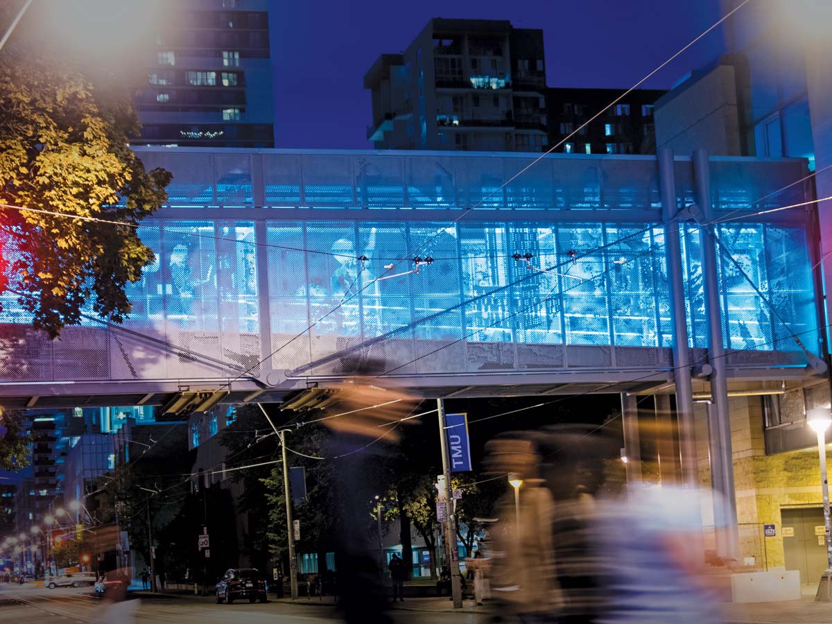 A glass walkway over a city street is lit with dim blue light. 