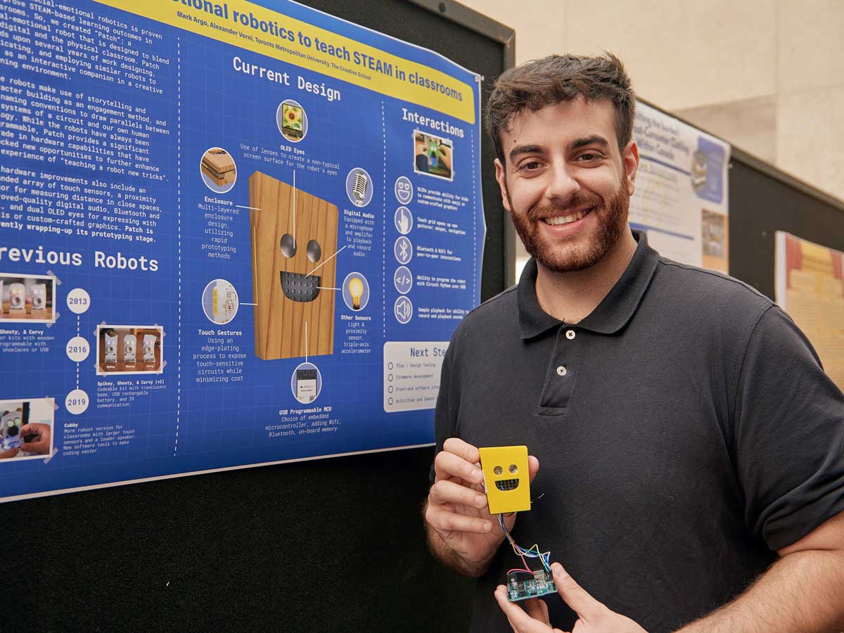 A man in a black shirt holds a small yellow robot with a happy face attached to a circuit board with his research poster in the background.