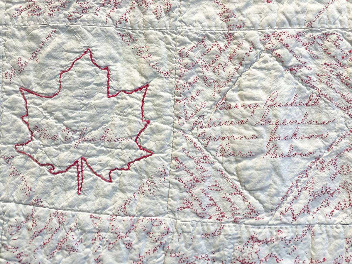 A quilt with a maple leaf and names embroidered in red thread on white fabric. 