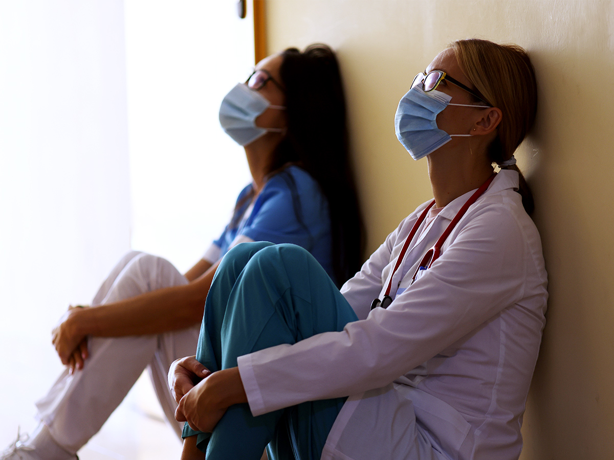 Two female healthcare workers, one in a blue uniform and the other in a light colour, sit against a wall with their arms around their legs. 