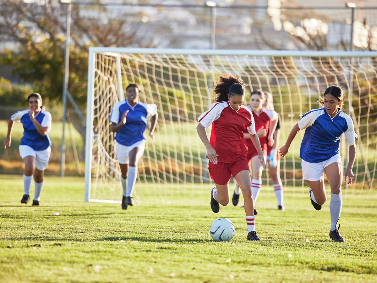 A group of teenage girls play soccer on an outdoor field. One team wears red uniforms, and the other wears blue. A girl on the red team is dribbling the ball away from her goal.