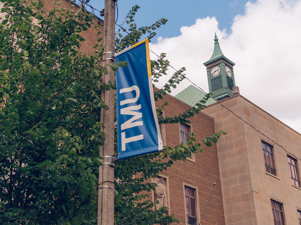 A blue university banner with “TMU” in white letters hangs on a pole outside of a brick campus building with a blue sky and clock tower in view