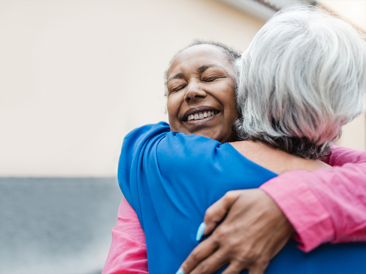 Two elderly women share a hug. The woman facing the camera has her eyes closed and is wearing a pink shirt. The woman facing away is wearing a blue shirt.