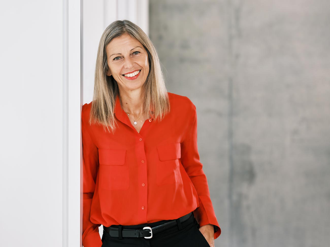 Professor Anna Triandafyllidou stands leaning against a white wall wearing a red shirt and black pants