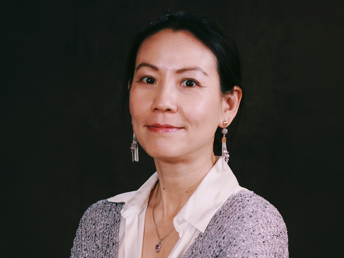 A headshot of professor Zhixi Zhuang, who looks at the camera against a dark background, wearing dangling earrings, a light grey sweater and a white-collared shirt