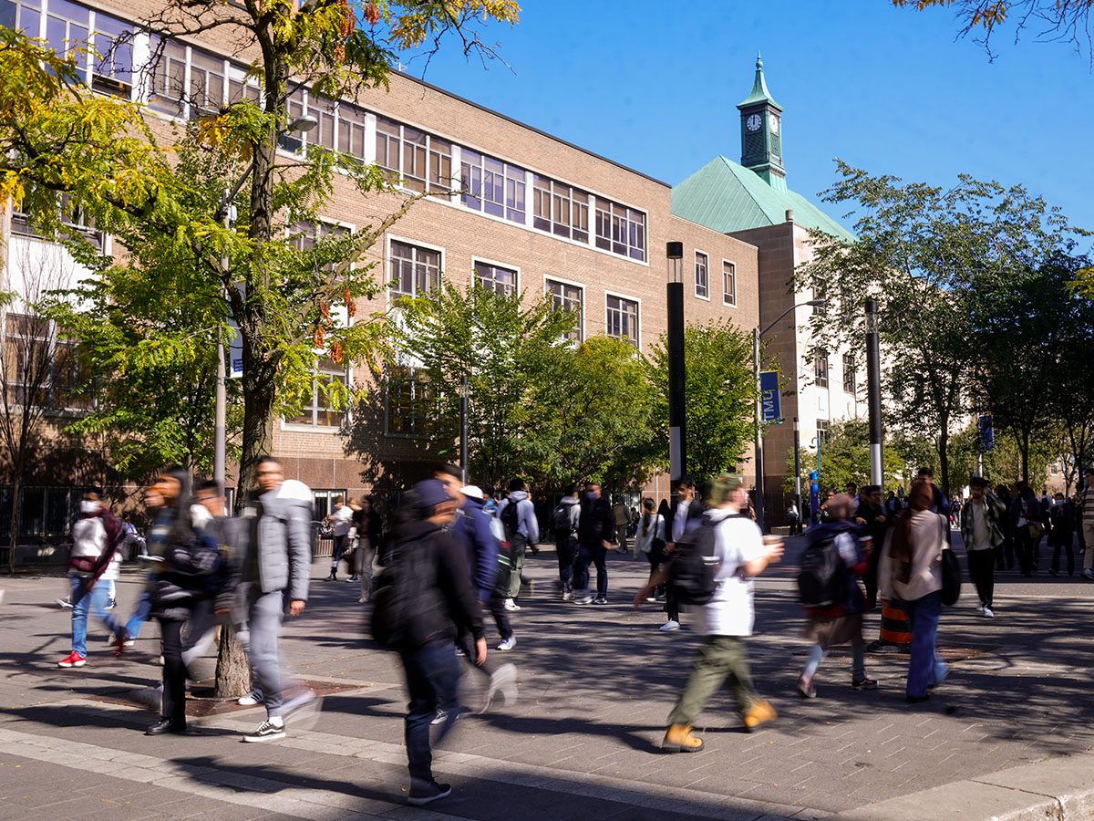 Students walk through TMU’s campus on a sunny day, with academic buildings and trees lining the pedestrian walkway. Motion blur captures the busy movement of people across campus.