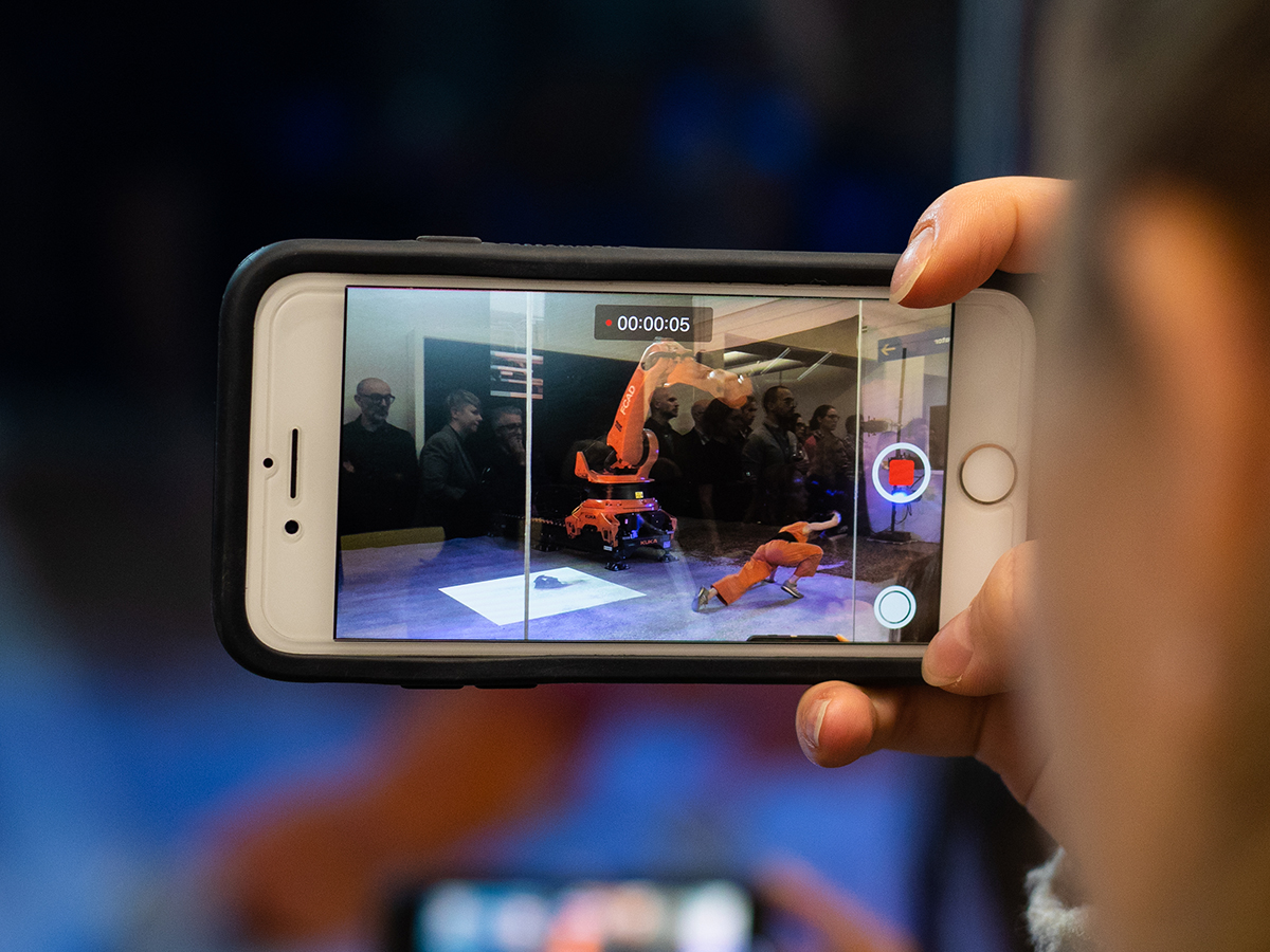 An onlooker records a performer dancing with a large mechanical arm with their smart phone