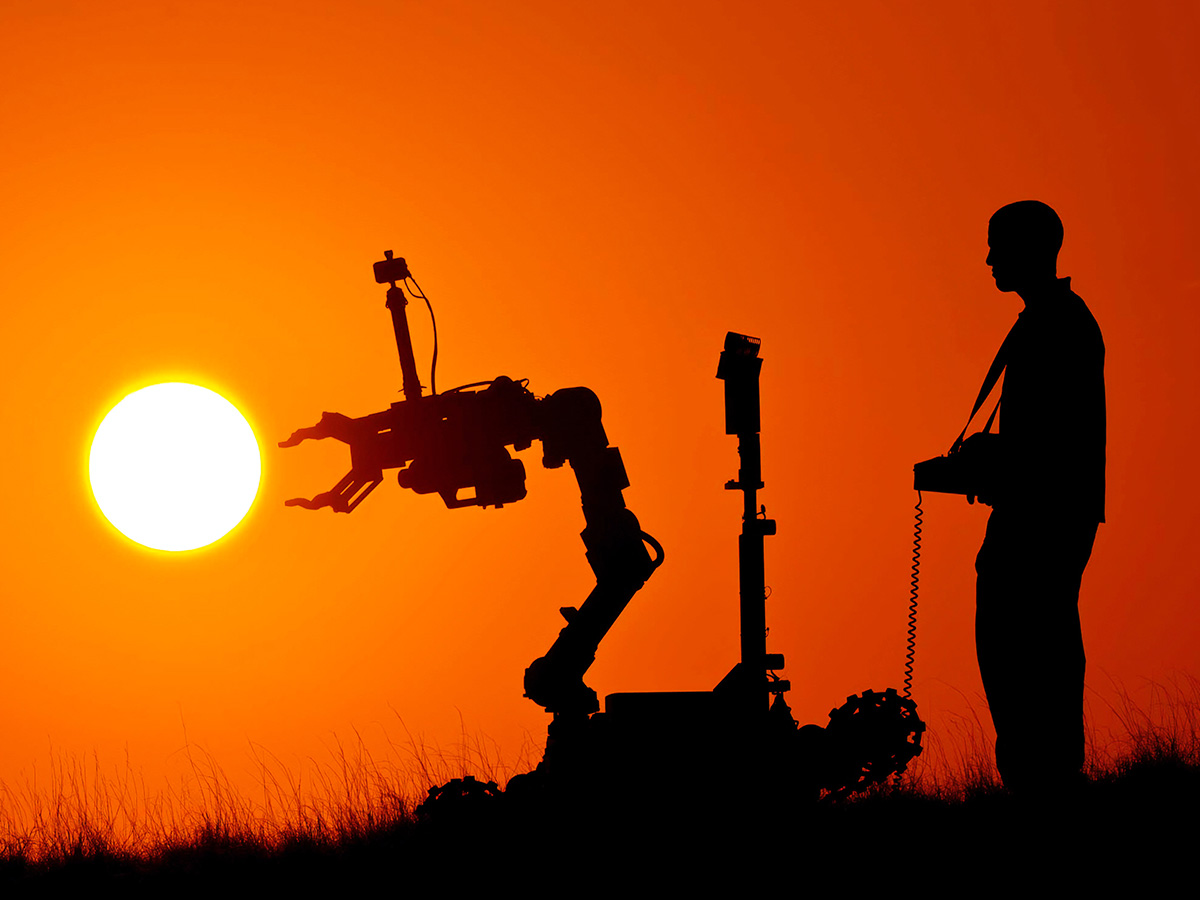 An individual controls a robot with a large robotic arm in a field at sunset