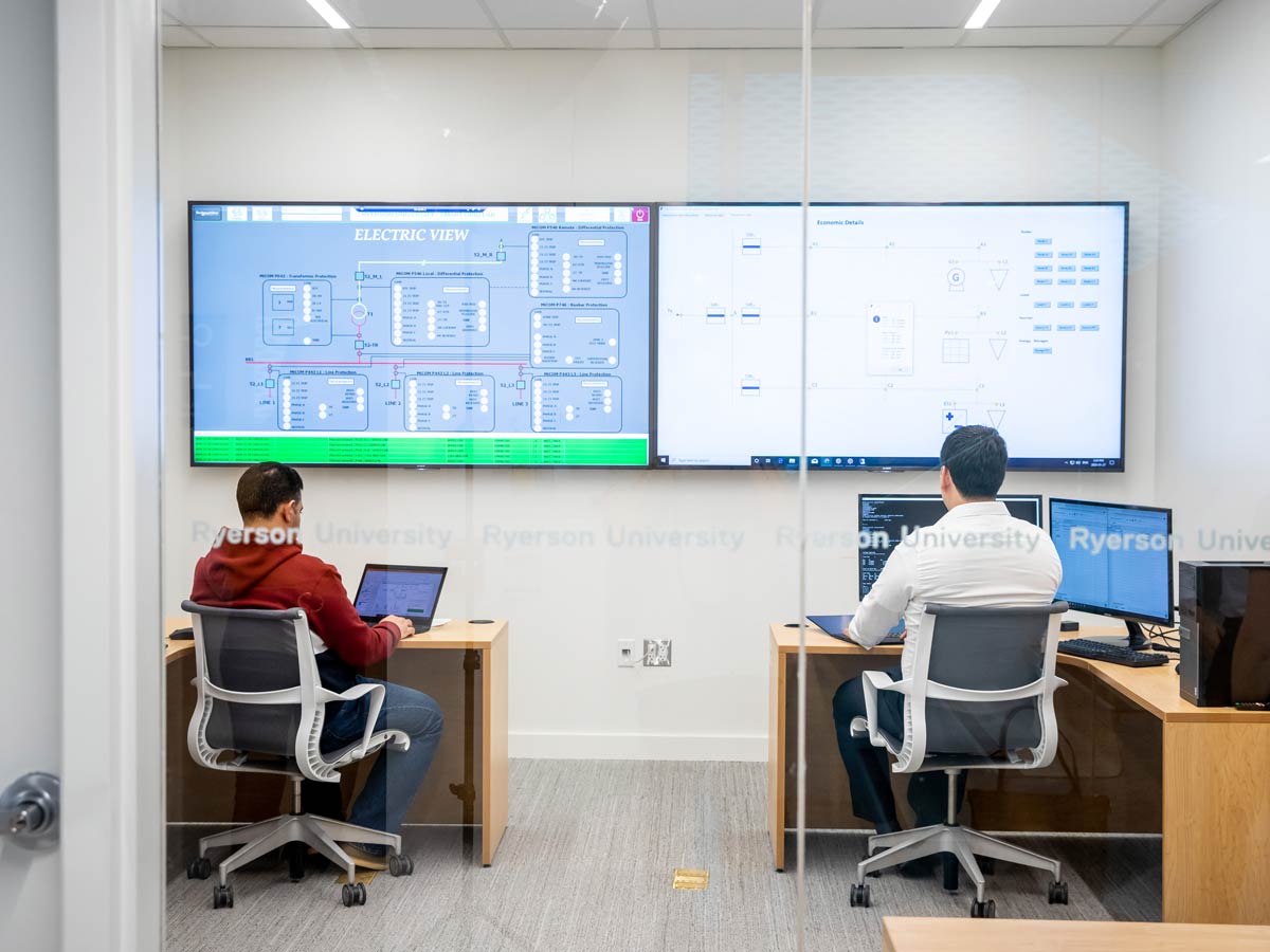 Two researchers work at computers in an office with two large monitors on the wall.