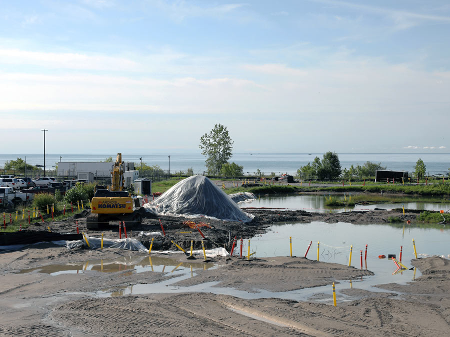 A front loader next to a mound of soil covered in a plastic tarp at the centre of a large tract of empty wetland.