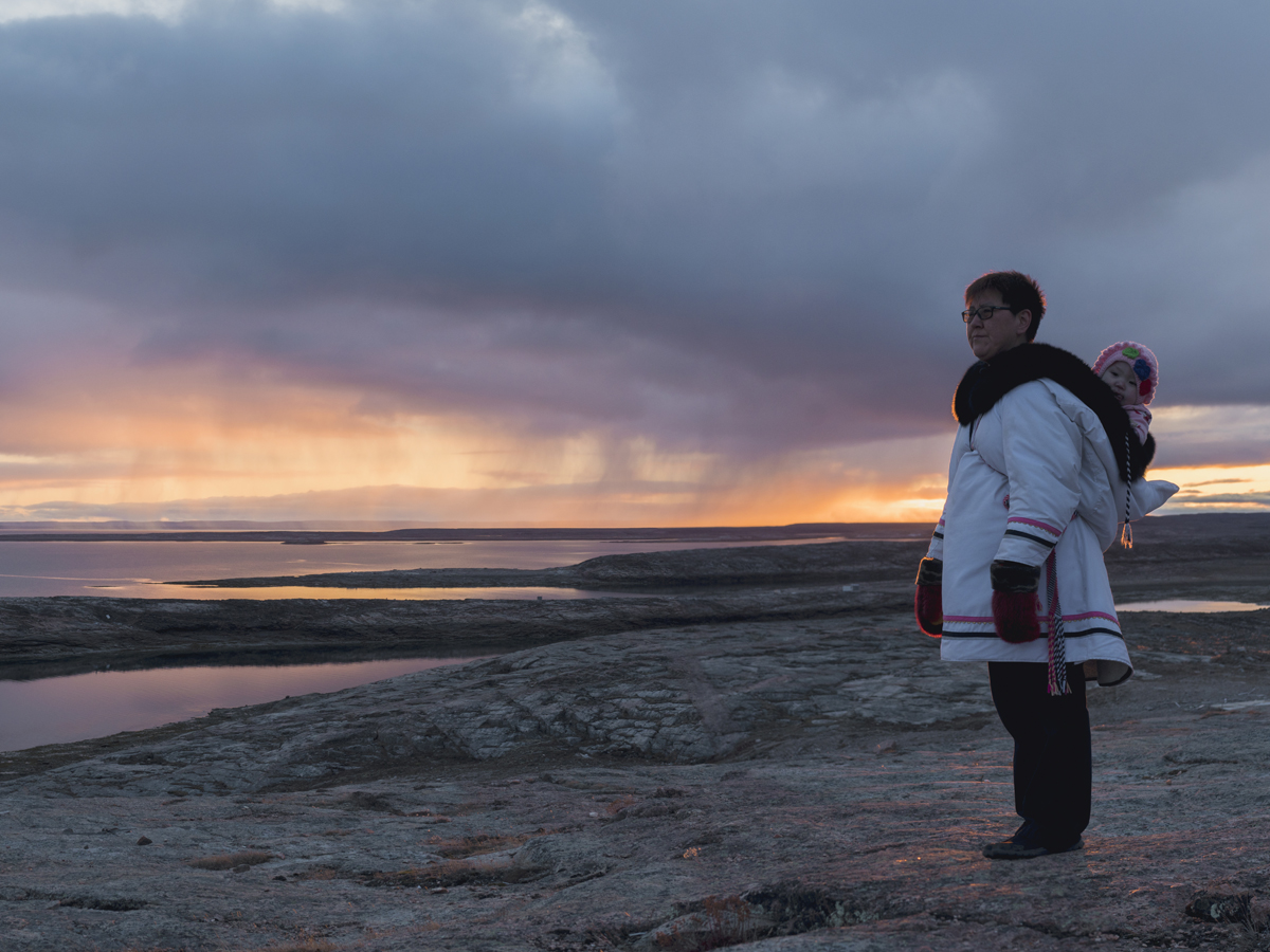 A woman with a baby on her back standing on a shoreline in Canada's north during the evening