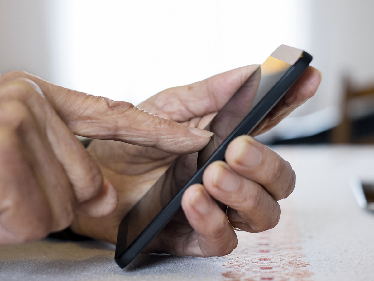 A pair of slender, wrinkled hands hold a smartphone, one finger touching the screen.