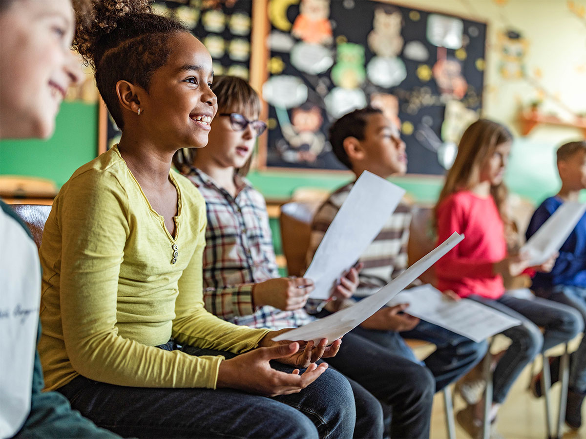 A group of children in a classroom are holding sheet music, singing and smiling.