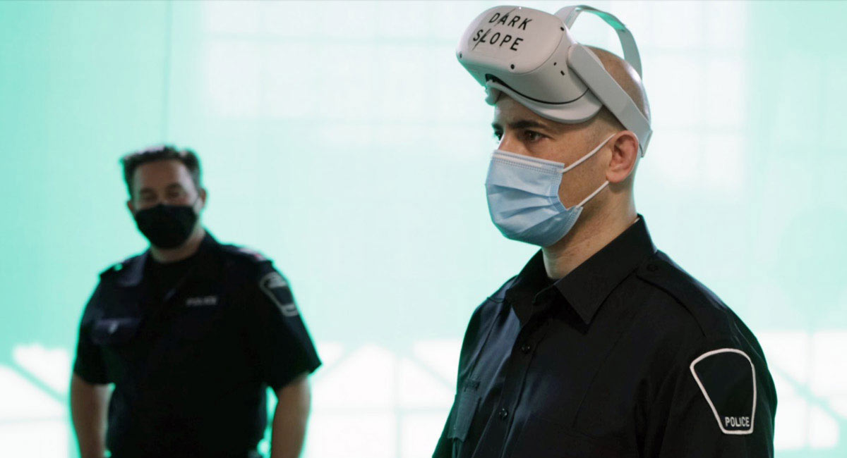 Two police officers stand in a room, both wearing face masks. One police officer wears a VR headset branded with a Dark Slope company logo.