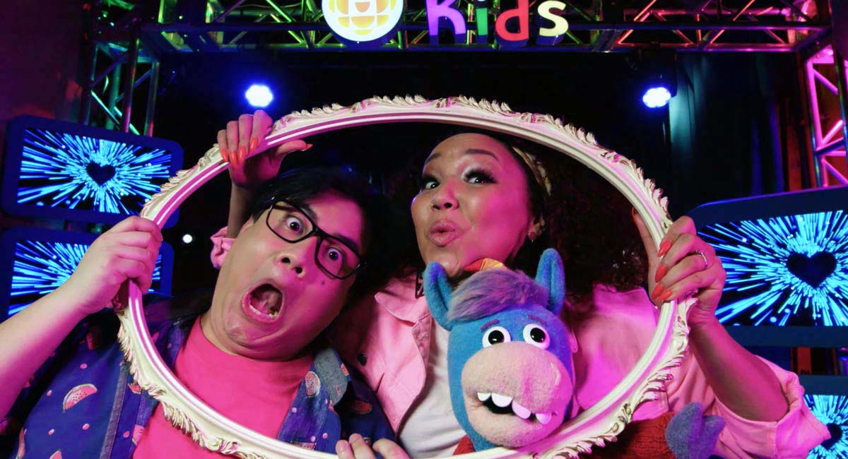 In a production studio, two people make funny faces along with a blue horse puppet while they look through an empty picture frame, creating the impression that they are the picture inside the frame.