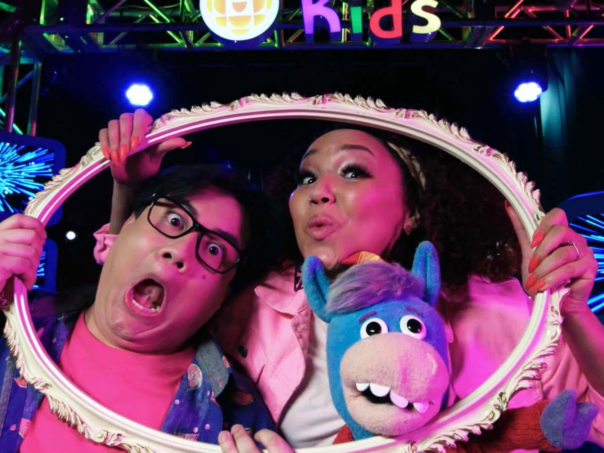 In a production studio, two people make funny faces along with a blue horse puppet while they look through an empty picture frame, creating the impression that they are the picture inside the frame.