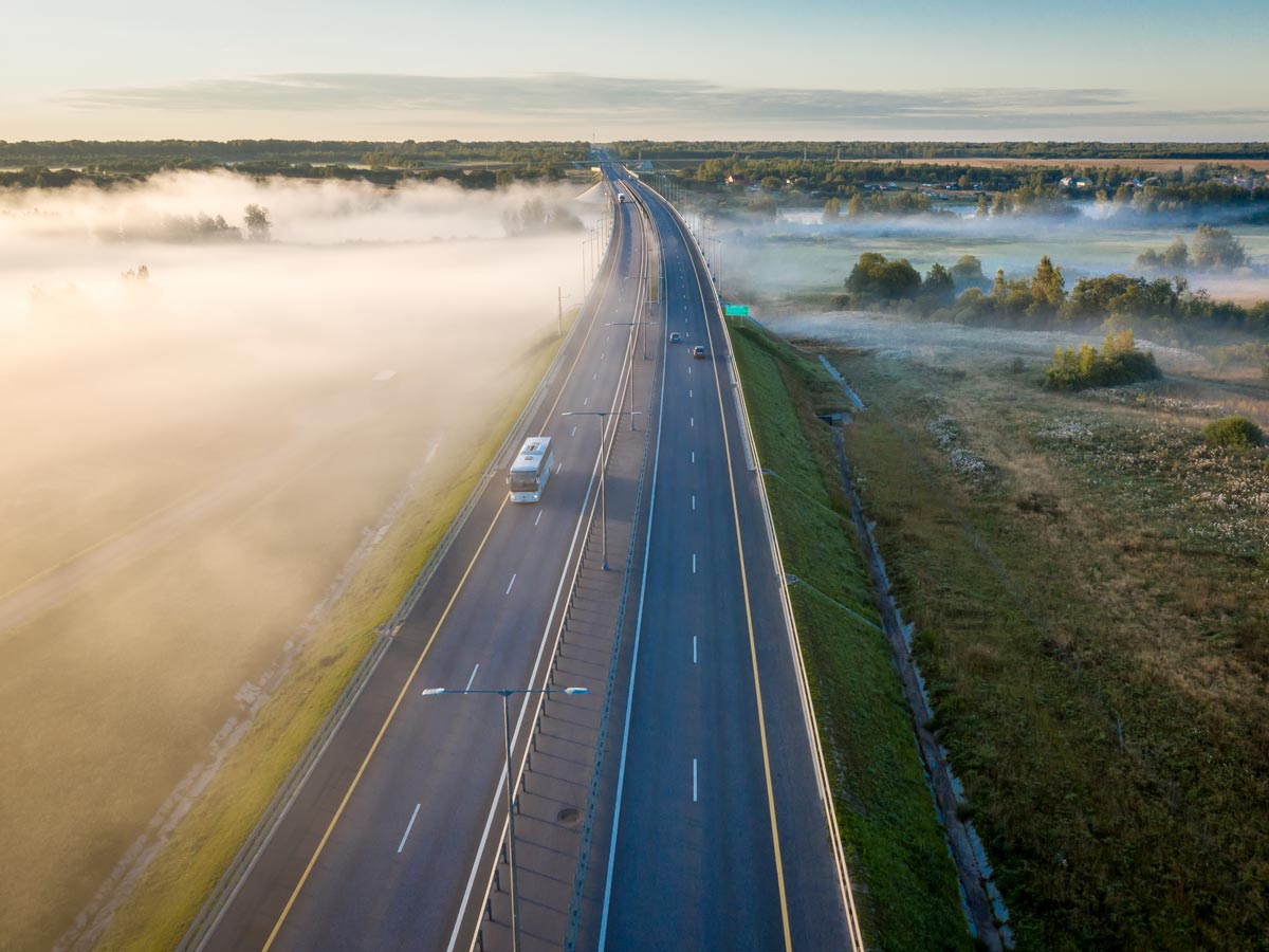 Aerial shot of a foggy highway with a few cars. There are open green fields to either side of the highway. Sparse housing can be seen in the distance.