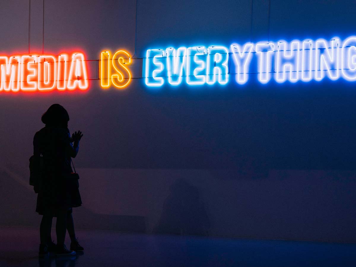 Two people stand closely together in a dark room, silhouetted by a neon sign above them that says "Media is Everything."
