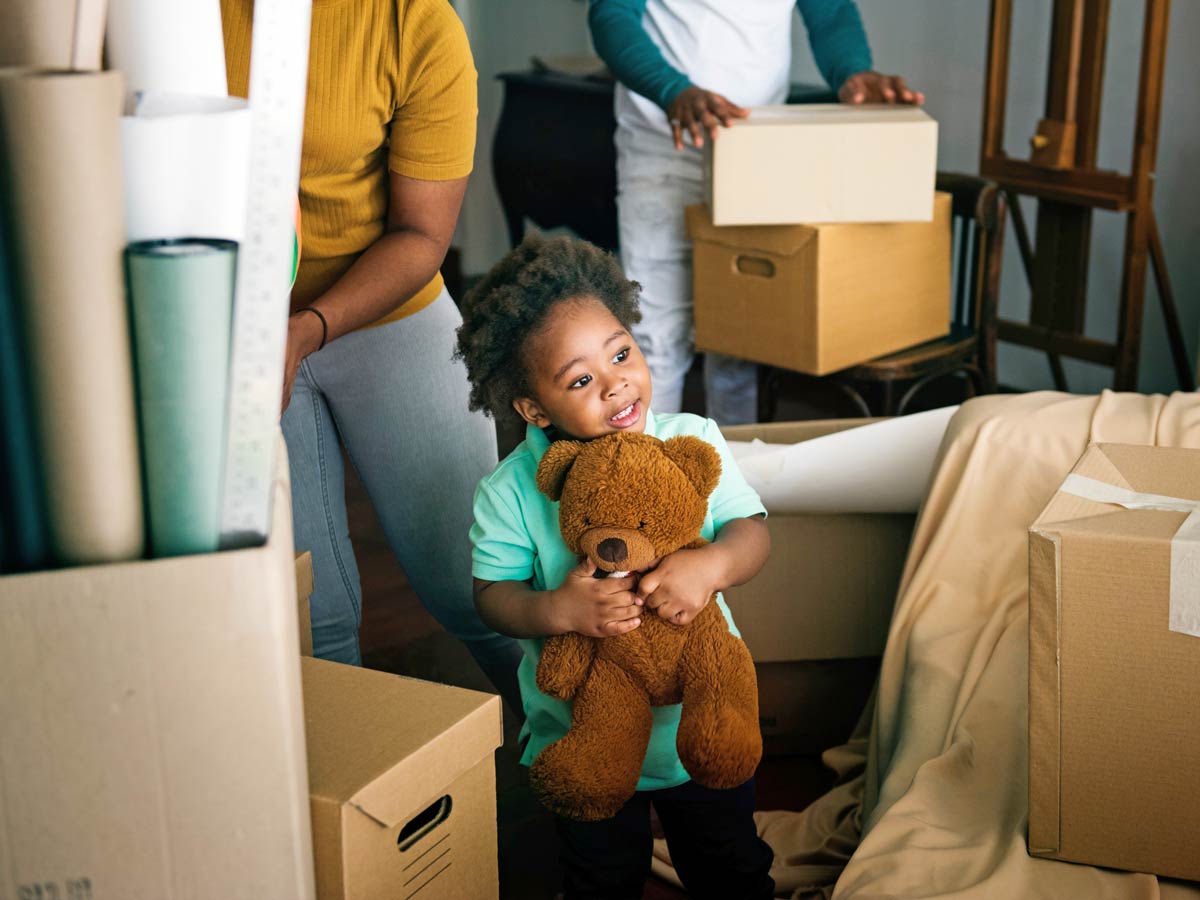 A small Black child holding a teddy bear is surrounded by boxes and adults packing in the background