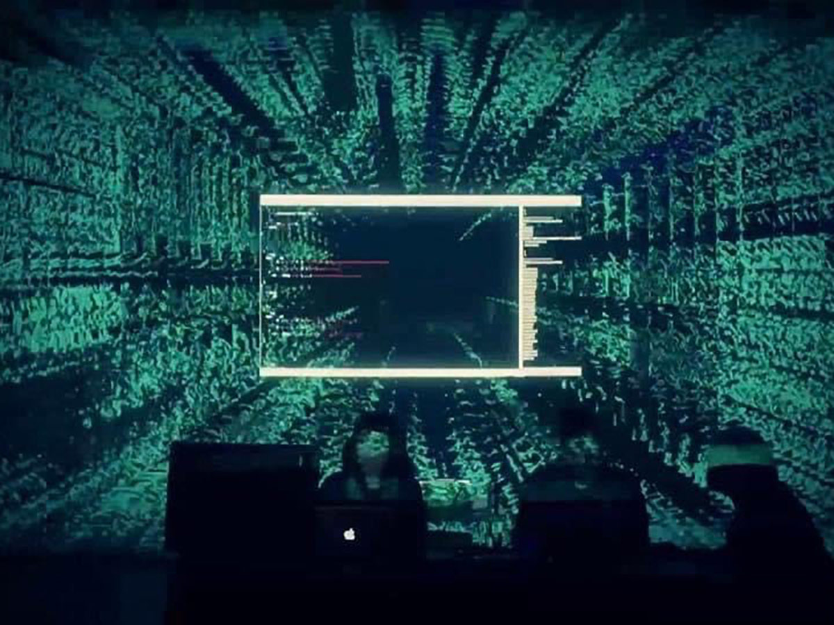 Three silhouetted DJs perform in a dark room with a projection of a glitchy computer screen showing lines of code on the wall behind them. 