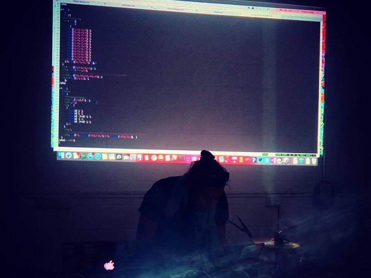 A silhouetted DJ performs in a dark room with a projection of a computer screen showing lines of code on the wall behind her.