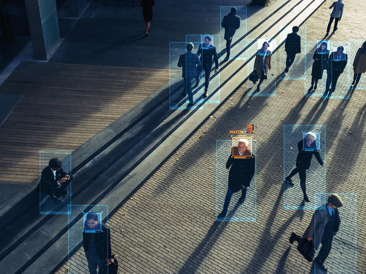 A view of an urban pedestrian walkway from the perspective of a security camera. The pedestrians are highlighted by transparent blue squares, and one man’s face is highlighted in orange with the word “match” next to it.