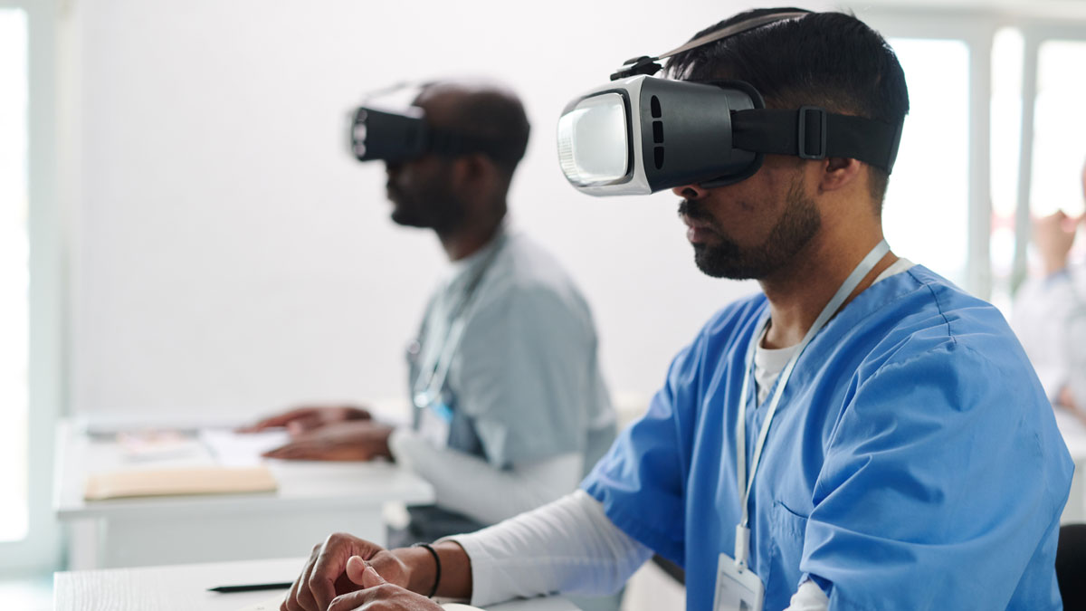 Two students wearing medical scrubs sit at desks wearing VR headsets. 