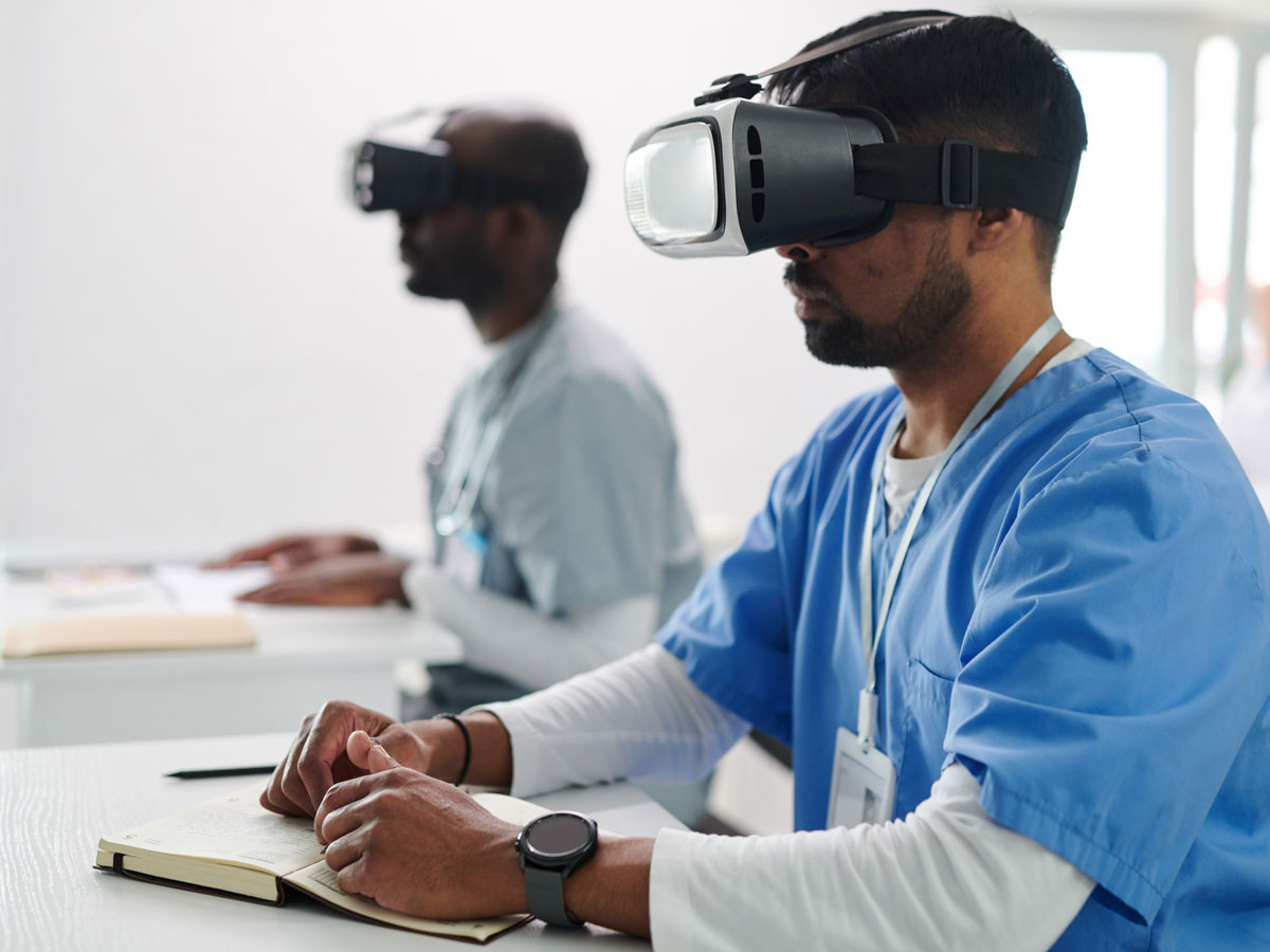 Two students wearing medical scrubs sit at desks wearing VR headsets. 