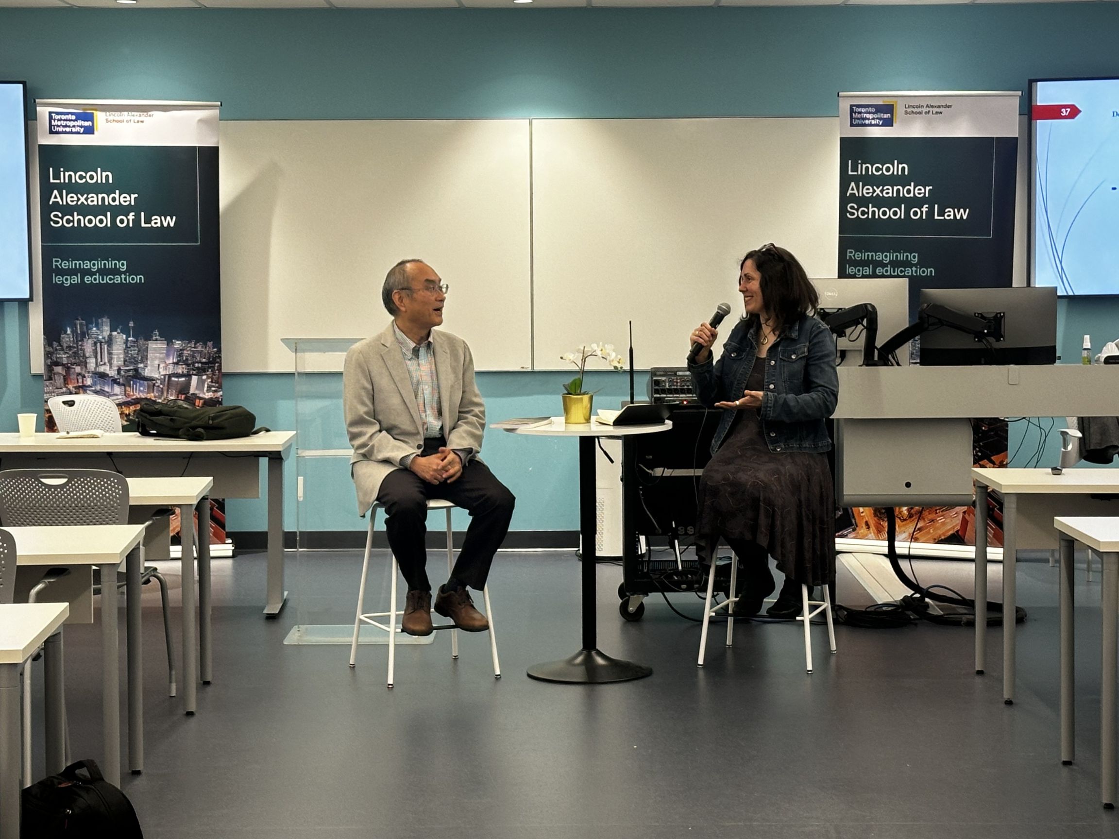 A man and a woman sitting in the middle of a classroom. The woman is holding a microphone and seems to be introducing the man. Banners of the Lincoln Alexander Law School are visible in the background,