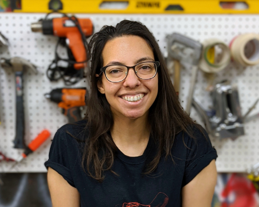 Mariana smiles at the camera. She is in front of the tool wall in The Workshop.
