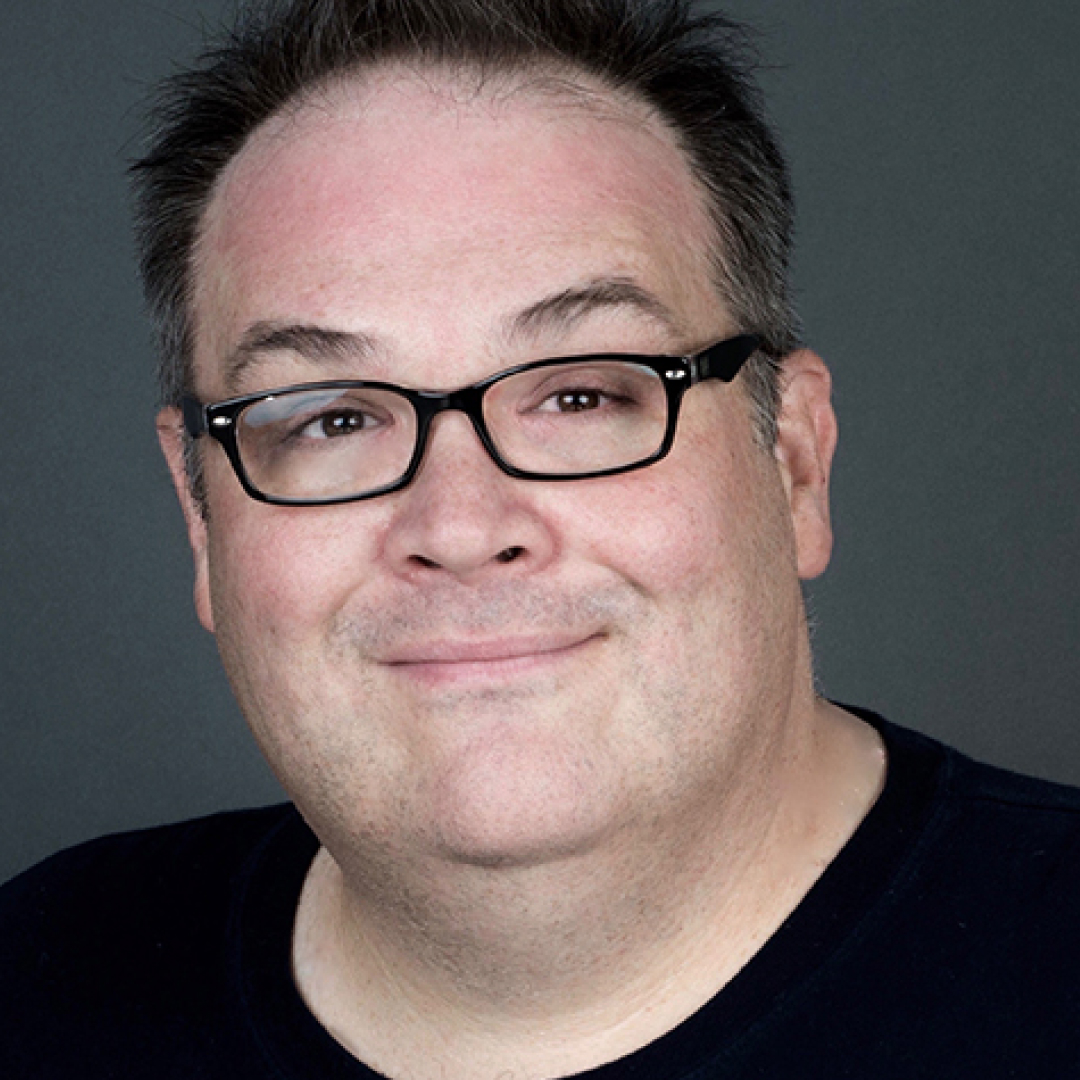 Close-up headshot of Denis McGrath smiling. He has short dark hair and glasses and is wearing a black shirt.