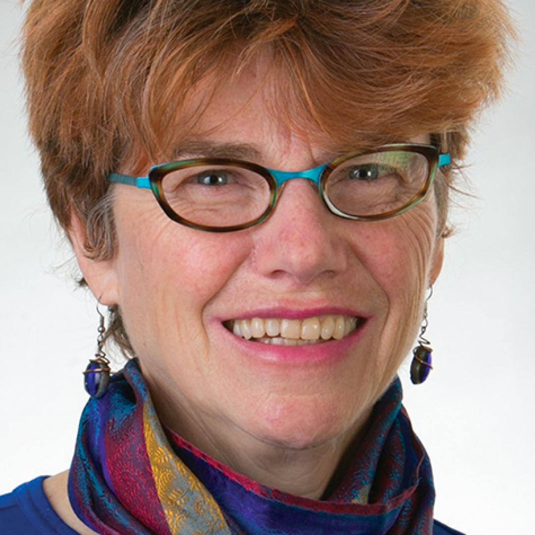 Close-up headshot of Dorothy Kidd smiling. She has short reddish hair, glasses, and is wearing a blue top with a multicoloured scarf.