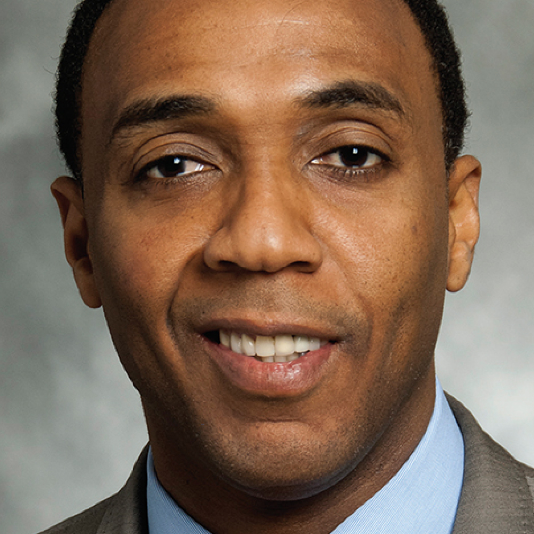 A close-up headshot of Dwight Drummond. He has short dark hair and is smiling slightly. He is wearing a grey suit jacket, light blue shirt, and tie, posed against a grey background.