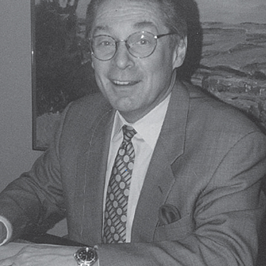 Black-and-white headshot of Jim Thompson smiling. He is wearing glasses, a suit, a patterned tie, and a watch, sitting at a desk.