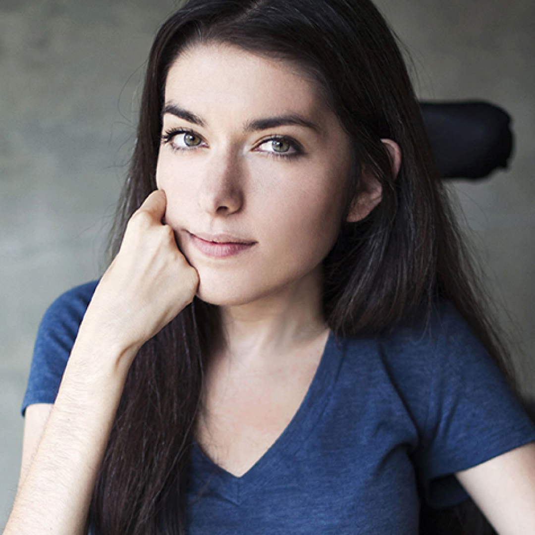 A close-up headshot of Maayan Ziv, resting her face on her hand, with long dark brown hair, wearing a blue T-shirt.