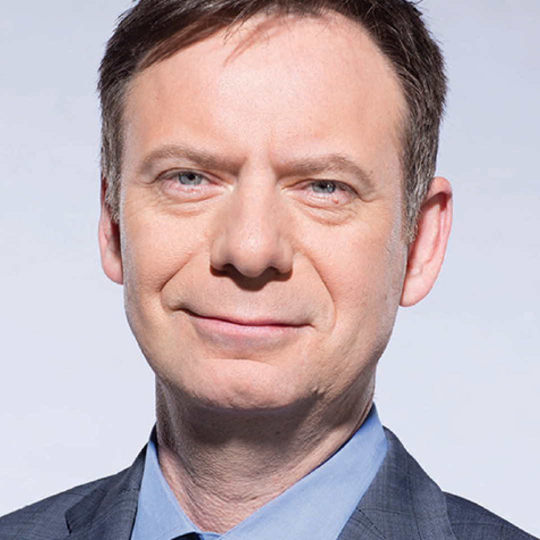 A close-up headshot of Mark Millier, with short brown hair, wearing a blue suit jacket and tie.